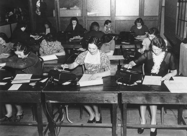 Black and white photograph. Twelve humans sit, quite packed, in rows at small desks. Each person has a large calculator in front of them. In terms of size and button shape they look like Comptometers, but they also seem to have rounded corners and cables leading from the back. Ten of the humans I would describe as white women, one I think is a white man, and the last one is a black man. The desks have enough space for the calculator, plus maybe two pieces of paper side by side without overlap.