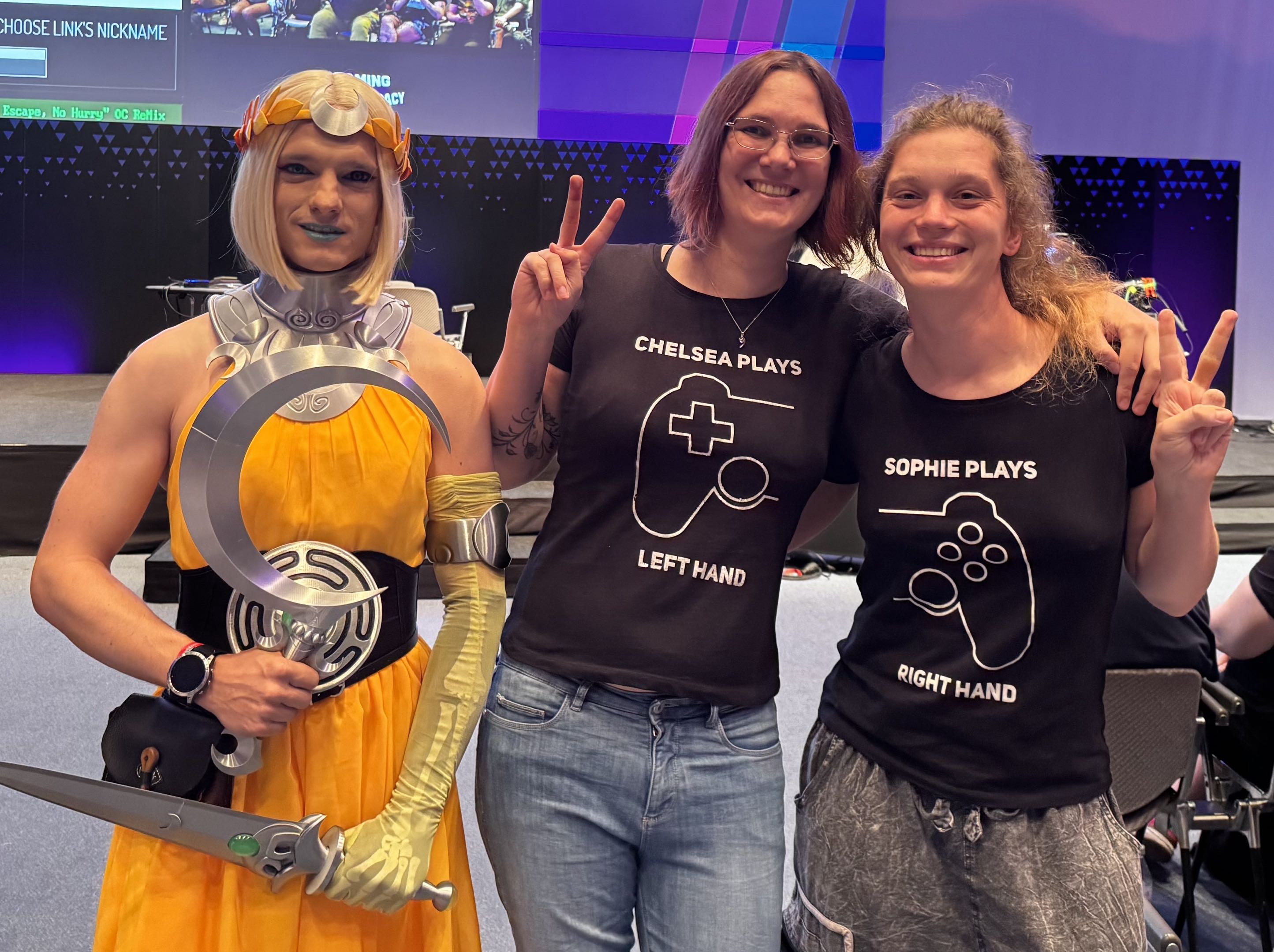 Chelsea and Sophie pose with a Melinoe cosplayer. Melineo is the main character in Hades II. Chelsea wears a shirt that says "Chelsea plays left hand" with the outline of the left side of a Playstation controller. Sophie wears a matching tshirt saying "Sophie players right hand".
